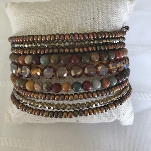 Könnte beinhalten: Ein Stapel von acht Armbändern aus braunen, goldenen und durchsichtigen Perlen. Die Armbänder sind in einem Kreis auf einer weißen Stoffoberfläche angeordnet.