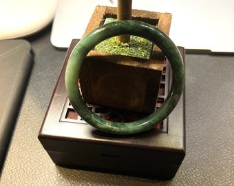 May include: A green jade bangle bracelet resting on a wooden box with a green moss lining. The bangle is smooth and polished.