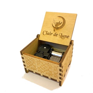 May include: A wooden music box with a carved design on the lid. The lid is open and the music box mechanism is visible. The lid has a fairy flying around a crescent moon with the text "Clair de Lune" engraved on it.
