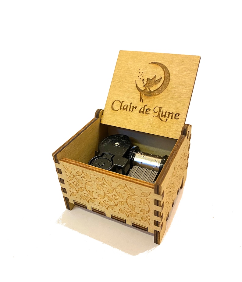 May include: A wooden music box with a carved design on the lid. The lid is open and the music box mechanism is visible. The lid has a fairy flying around a crescent moon with the text "Clair de Lune" engraved on it.