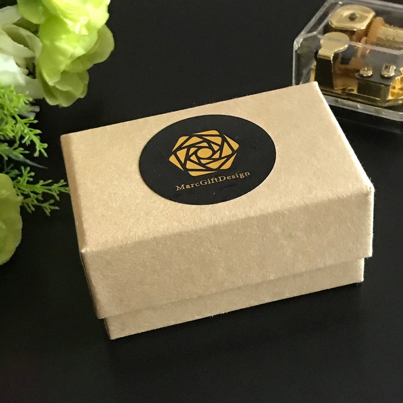 Op de afbeelding: Een bruine kartonnen doos met een zwarte en gouden sticker waarop "MarcGiftDesign" staat. De sticker heeft een gestileerd rozenmotief.