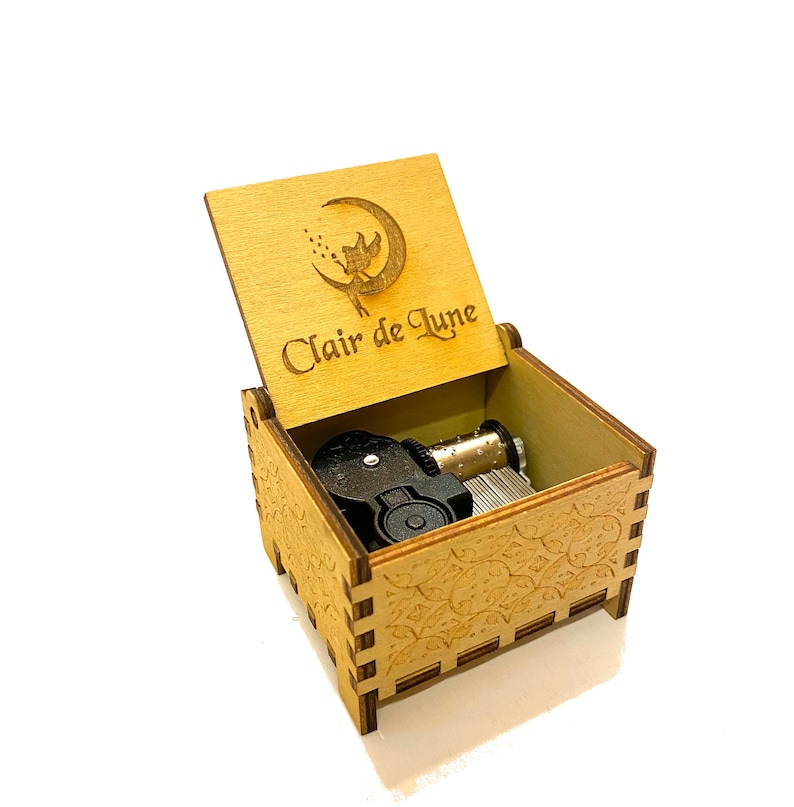 May include: A wooden music box with a carved design of a fairy on a crescent moon. The lid is open and the inscription "Clair de Lune" is visible. The music box has a black winding mechanism.