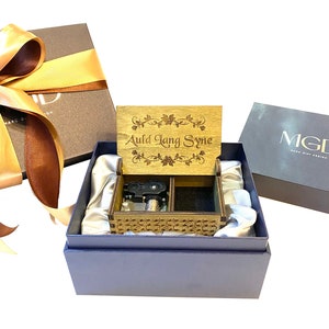 Puede incluir: Una caja de música de madera con un diseño floral y la inscripción "Auld Lang Syne". La caja de música está abierta y el mecanismo es visible. La caja de música está en una caja de regalo azul con un forro de satén blanco.