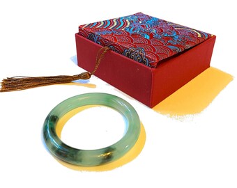 May include: A jade bangle bracelet with green and black hues rests beside a red box with a patterned lid. The box has a tassel attached. The bracelet is a circular shape, and the box is rectangular.