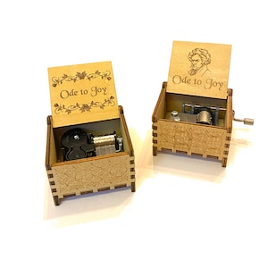 May include: Two wooden music boxes with the inscription "Ode to Joy" on the lid. One box has a floral design and the other has a portrait of a man. Both boxes have a winding mechanism on the side.