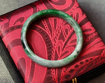 May include: A green jade bangle bracelet with a smooth, polished finish. The bracelet is displayed on a red velvet cushion with a floral pattern.