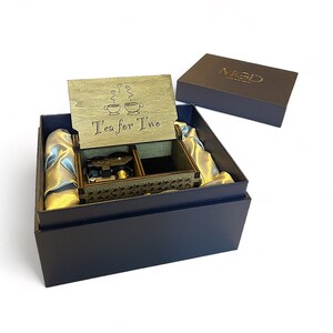 May include: A wooden music box with a lid that says "Tea for Two" and features a design of two teacups. The music box is in a blue box with a satin lining.