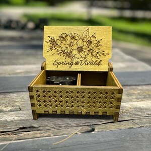 May include: A wooden music box with a floral design on the lid. The lid is open and the music box plays the song "Spring" by Vivaldi. The music box is made of light brown wood and has a delicate, intricate design.