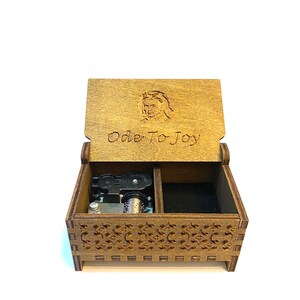 Op de afbeelding: Een houten muziekdoos met een open deksel, met de tekst "Ode To Joy" en een gesneden afbeelding. De doos heeft een ingewikkeld geometrisch ontwerp aan de voorkant en bevat een mechanisch muziekmechanisme. De binnenkant is gevoerd met zwarte stof.