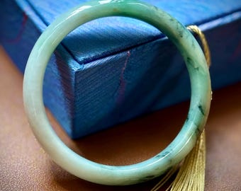 May include: A jade bangle bracelet with a gradient of light green to white. The bracelet is smooth and polished, with a slight translucence. A gold tassel hangs near the bracelet. The background includes a blue box and a brown surface.