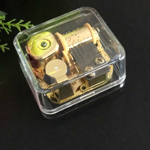 Op de afbeelding: Een gouden muziekdoosmechanisme in een doorzichtige plastic behuizing. Het mechanisme heeft een opwindsleutel en een kleine, ronde, gele knop.