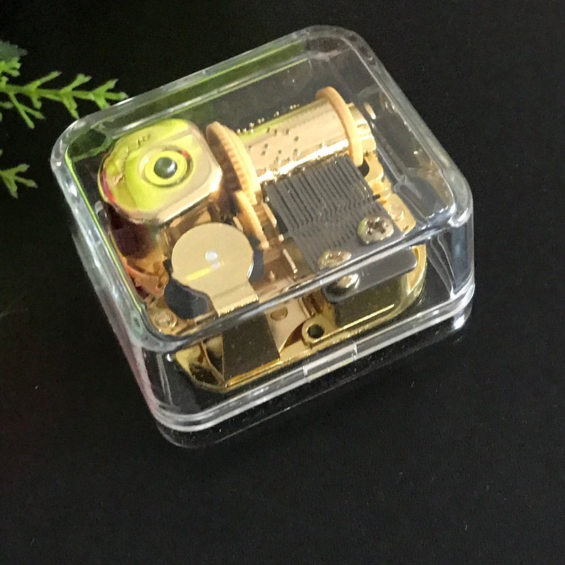 Op de afbeelding: Een gouden muziekdoosmechanisme in een doorzichtige plastic behuizing. Het mechanisme heeft een opwindsleutel en een kleine, ronde, gele knop.