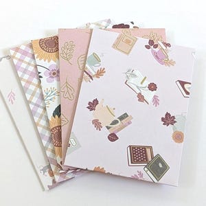 May include: A set of five rectangular greeting cards arranged in a fan shape. The designs feature various patterns including autumn leaves, sunflowers, plaid, and illustrations of books, teapots, and mugs in shades of pink, brown, and cream.