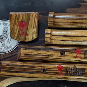 Może przedstawiać: Zestaw drewnianych akcesori&oacute;w do grillowania z czerwonym logo Weber. Zestaw zawiera otwieracz do butelek, monetę z kogutem i kilka prostokątnych element&oacute;w. Drewno ma wz&oacute;r w paski.