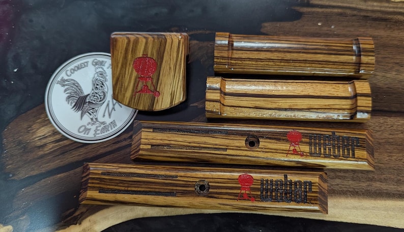 Może przedstawiać: Zestaw drewnianych akcesori&oacute;w do grillowania z czerwonym logo Weber. Zestaw zawiera otwieracz do butelek, monetę z kogutem i kilka prostokątnych element&oacute;w. Drewno ma wz&oacute;r w paski.