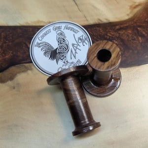 May include: Two dark brown wooden grill handles with a circular design. A white sticker with a rooster illustration and the text "The Coolest Grill Handles on Earth ZAGA" is also present. The handles have a cylindrical shape with a flared base and top.