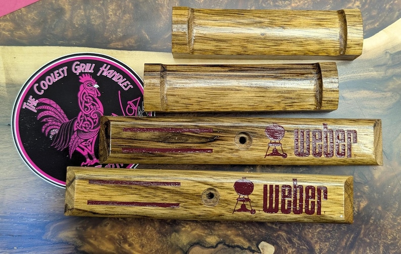 Puede incluir: Mangos de parrilla de madera con un acabado de vetas de madera natural. Dos mangos est&aacute;n grabados con la palabra "Weber" y un gr&aacute;fico de parrilla en rojo. Tambi&eacute;n hay una pegatina circular con un gr&aacute;fico de gallo y el texto "The Coolest Grill Handles".