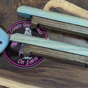 Puede incluir: Asas de parrilla de madera con acabado azul claro y base marr&oacute;n oscuro. Un objeto cuadrado azul claro con un gr&aacute;fico de parrilla negro y una pegatina redonda con el texto "Coolest Grill on Earth" tambi&eacute;n est&aacute;n en la imagen.