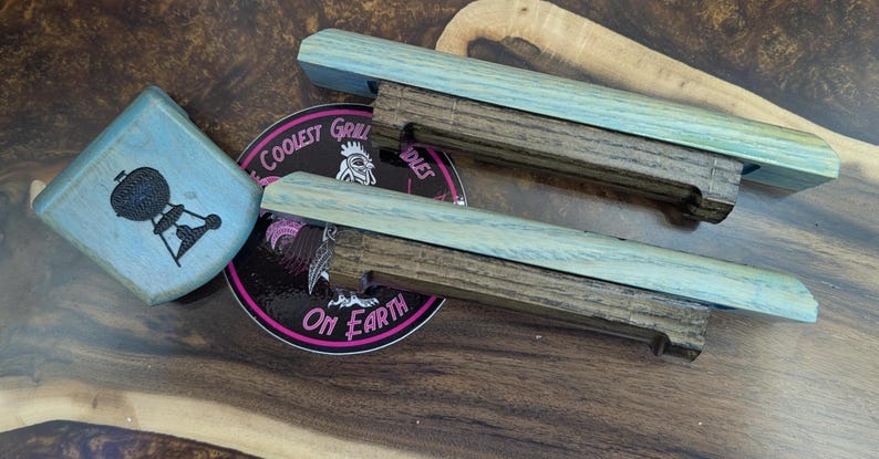 Puede incluir: Asas de parrilla de madera con acabado azul claro y base marr&oacute;n oscuro. Un objeto cuadrado azul claro con un gr&aacute;fico de parrilla negro y una pegatina redonda con el texto "Coolest Grill on Earth" tambi&eacute;n est&aacute;n en la imagen.