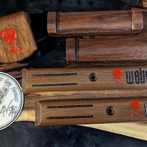 May include: Wooden grill handle replacements and a square wooden piece with a red design. Two long wooden pieces have the word "Weber" in black with a red accent. A sticker with a rooster and text is also present.