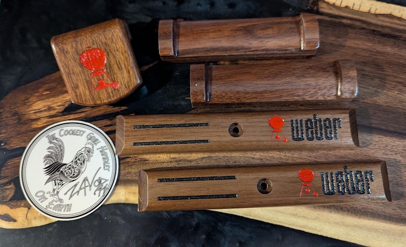 May include: Wooden grill handle replacements and a square wooden piece with a red design. Two long wooden pieces have the word "Weber" in black with a red accent. A sticker with a rooster and text is also present.
