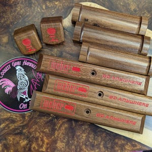 Op de afbeelding: Houten grill handgreep vervangingen en kubussen met rode Weber logo's en tekst. De handgrepen zijn rechthoekig met een gat voor montage. Een sticker met een haan en de tekst "Coolest Grill Handles On" is ook aanwezig.