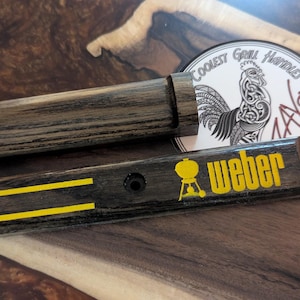 Könnte beinhalten: Ein dunkles Grillgriff-Set aus Holz. Das Set enthält einen Griff, ein kleines rechteckiges Stück mit dem Wort "Weber" in Gelb und einen runden Aufkleber mit einer Hahn-Illustration und dem Text "Coolest Grill Handles".