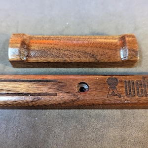 May include: Two wooden barbecue accessories. The top piece is a small, rectangular rest. The bottom piece is a rectangular base with the word "Weber" and a grill logo etched into the wood. Both pieces are made of dark wood.