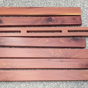 Peut inclure: Vue aérienne de plusieurs morceaux de bois de cèdre non finis. Il y a sept pièces rectangulaires, dont une plus longue avec trois trous espacés. Le bois est brun rougeâtre et présente un veinage visible.