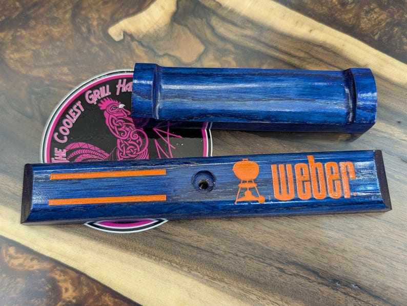 May include: Two blue wooden grill accessories. One is rectangular with the word "Weber" in orange, and the other is a cylindrical handle. A sticker with a rooster is visible.
