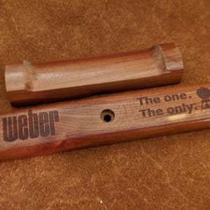 Pode incluir: Dois acess&oacute;rios de churrasco em madeira. Um &eacute; uma pe&ccedil;a retangular com a palavra "Weber" gravada, juntamente com a frase "The one. The only." e um logotipo de grelhador. O outro &eacute; uma pe&ccedil;a de madeira curva. Ambos s&atilde;o de cor castanho quente.