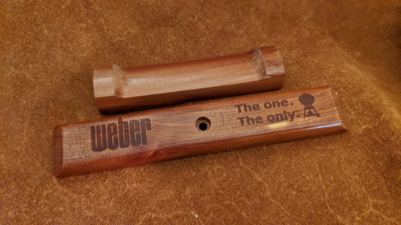 Pode incluir: Dois acess&oacute;rios de churrasco em madeira. Um &eacute; uma pe&ccedil;a retangular com a palavra "Weber" gravada, juntamente com a frase "The one. The only." e um logotipo de grelhador. O outro &eacute; uma pe&ccedil;a de madeira curva. Ambos s&atilde;o de cor castanho quente.