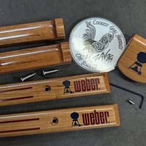 Może przedstawiać: Drewniane zamienniki uchwytów do grilla i akcesoria. Zawiera dwa elementy uchwytu, dwa elementy uchwytu do grilla Weber z logo Weber, okrągłą odznakę z kogutem i małą drewnianą szpatułkę z logo grilla.