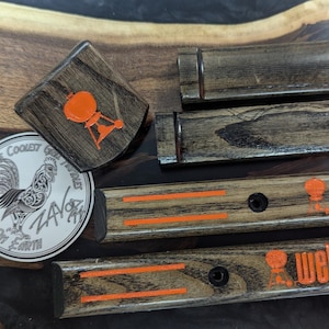 Puede incluir: Colecci&oacute;n de accesorios de parrilla de madera, que incluye una peque&ntilde;a pieza cuadrada con un logotipo de parrilla naranja, piezas rectangulares con rayas naranjas y la palabra "Weber", y una pegatina redonda con un gallo.