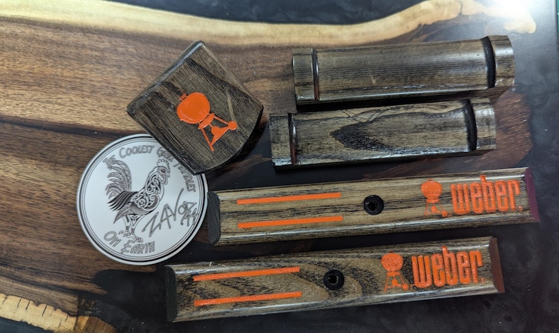 Puede incluir: Colecci&oacute;n de accesorios de parrilla de madera, que incluye una peque&ntilde;a pieza cuadrada con un logotipo de parrilla naranja, piezas rectangulares con rayas naranjas y la palabra "Weber", y una pegatina redonda con un gallo.