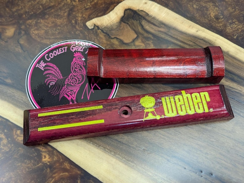 May include: A set of two wooden grill accessories with a dark red finish. One piece has the text "Weber" in yellow, and the other has a circular sticker with a rooster graphic and the text "The Coolest Grill".