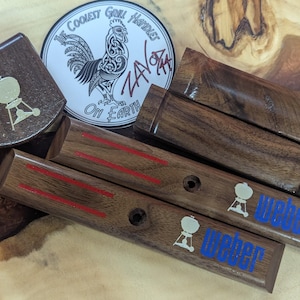 May include: Dark wooden grill handle replacements with red stripes and the word "Weber" in blue. A sticker featuring a rooster and the text "The Coolest Grill Handles On Earth" is also visible. The handles are made of dark wood.