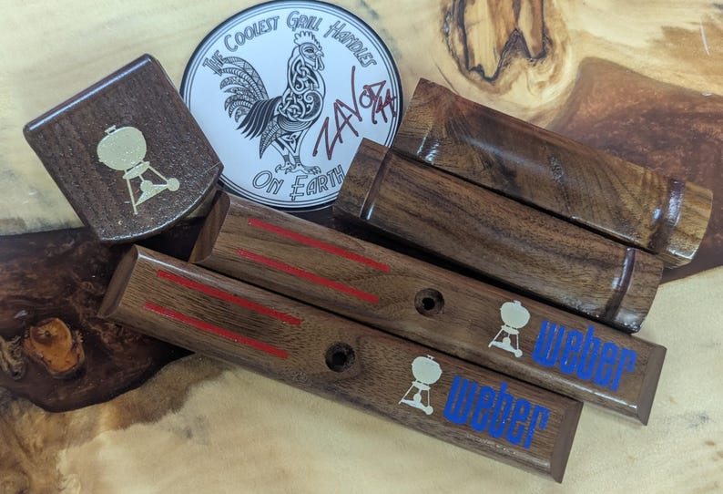 May include: Dark wooden grill handle replacements with red stripes and the word "Weber" in blue. A sticker featuring a rooster and the text "The Coolest Grill Handles On Earth" is also visible. The handles are made of dark wood.