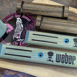 Puede incluir: Accesorios de parrilla de madera, incluyendo manijas y una pieza de esquina, se muestran sobre una superficie de madera. Las manijas son de color azul claro con detalles negros y el logotipo de Weber. Tambi&eacute;n se ve una pegatina con un gallo y texto.
