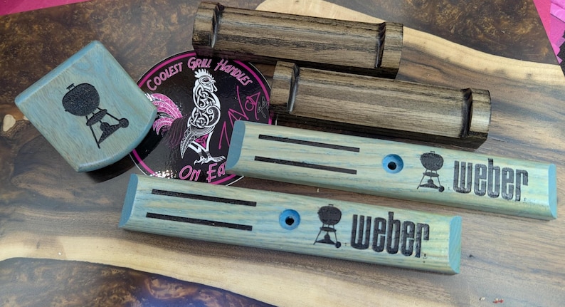 Puede incluir: Accesorios de parrilla de madera, incluyendo manijas y una pieza de esquina, se muestran sobre una superficie de madera. Las manijas son de color azul claro con detalles negros y el logotipo de Weber. Tambi&eacute;n se ve una pegatina con un gallo y texto.