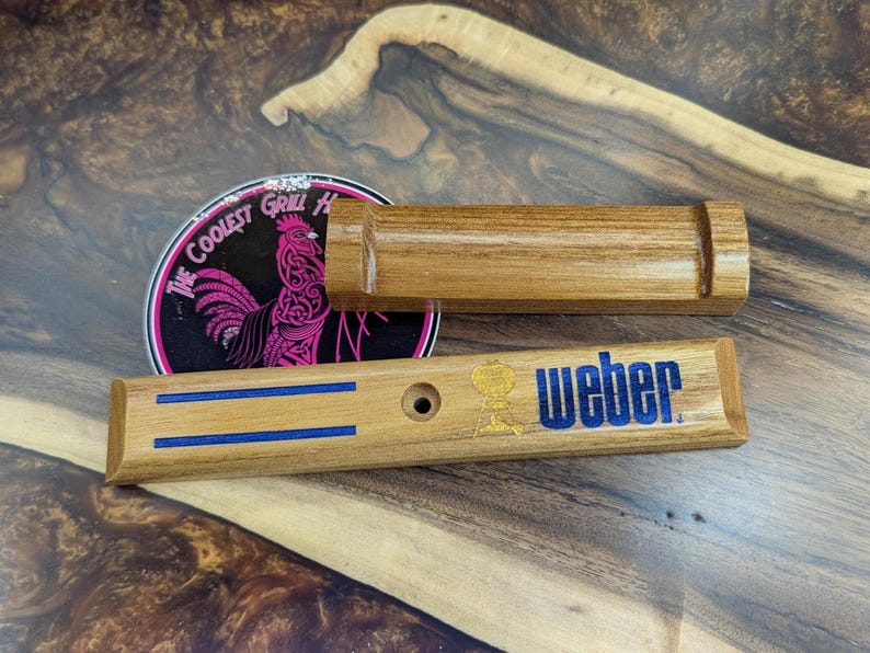 May include: Wooden grill handle and tool rest with the Weber logo in blue and gold. The handle has two blue stripes. A coaster with a rooster graphic and the text "The Coolest Grill House" is also visible.