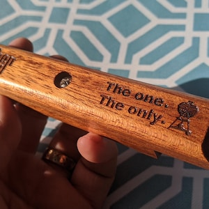 Pode incluir: Um abridor de garrafas de madeira com o logotipo "Weber" e o texto "The one. The only." &eacute; segurado. O abridor tem um pequeno orif&iacute;cio e um gr&aacute;fico de grelha. A madeira tem um gr&atilde;o natural.