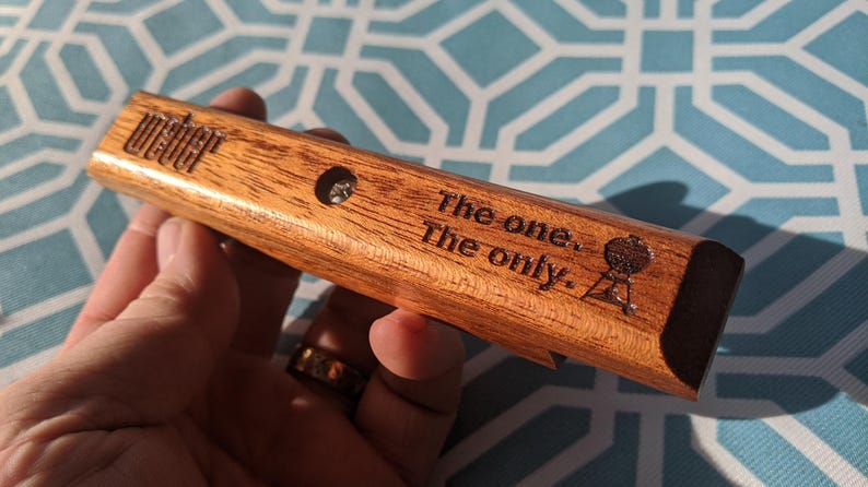 Pode incluir: Um abridor de garrafas de madeira com o logotipo "Weber" e o texto "The one. The only." &eacute; segurado. O abridor tem um pequeno orif&iacute;cio e um gr&aacute;fico de grelha. A madeira tem um gr&atilde;o natural.
