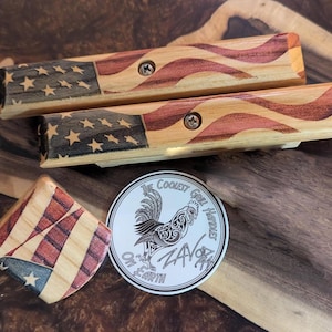 May include: Wooden grilling tools with an American flag design. The set includes two long tools and a small square piece, all featuring red, white, and blue wood grain patterns. A round sticker with a rooster graphic and the text "The Coolest Grill Hustler" is also present.