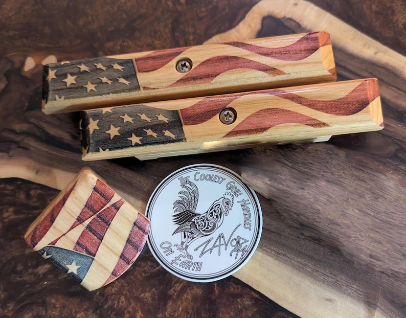 May include: Wooden grilling tools with an American flag design. The set includes two long tools and a small square piece, all featuring red, white, and blue wood grain patterns. A round sticker with a rooster graphic and the text "The Coolest Grill Hustler" is also present.