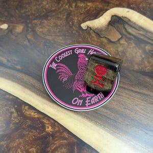 Puede incluir: Un cubo de madera oscura con un logotipo de parrilla rojo descansa sobre una pegatina negra y rosa. La pegatina dice "The Coolest Grill House On Earth" y presenta una ilustración de gallo rosa. El cubo tiene un acabado marrón oscuro.