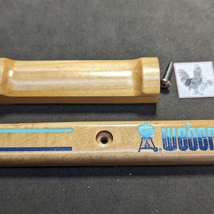 May include: Wooden Weber grill handle and decorative panel with blue and teal accents. The panel has the Weber logo and text. Includes a screw and a small square image of a rooster.