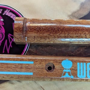 May include: Wooden grill handle and tool rest with the word "Weber" in turquoise. A circular sticker with a pink rooster and the text "The Coolest Grill Hands NA" is also visible. The items are made of wood.