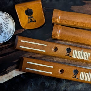 Puede incluir: Un conjunto de accesorios de parrilla de madera, que incluyen mangos y un plato peque&ntilde;o, con el logotipo de Weber y un emblema de gallo. Los art&iacute;culos est&aacute;n hechos de madera de tonos c&aacute;lidos y est&aacute;n dispuestos sobre una superficie oscura y texturizada.