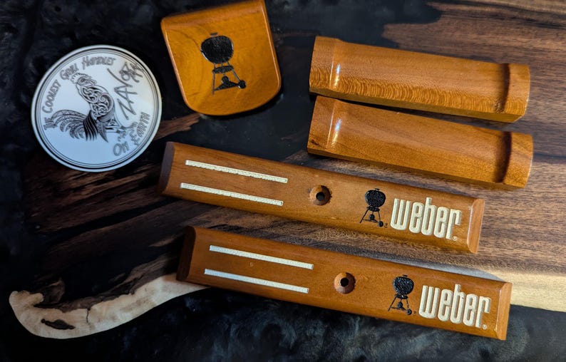 Puede incluir: Un conjunto de accesorios de parrilla de madera, que incluyen mangos y un plato peque&ntilde;o, con el logotipo de Weber y un emblema de gallo. Los art&iacute;culos est&aacute;n hechos de madera de tonos c&aacute;lidos y est&aacute;n dispuestos sobre una superficie oscura y texturizada.